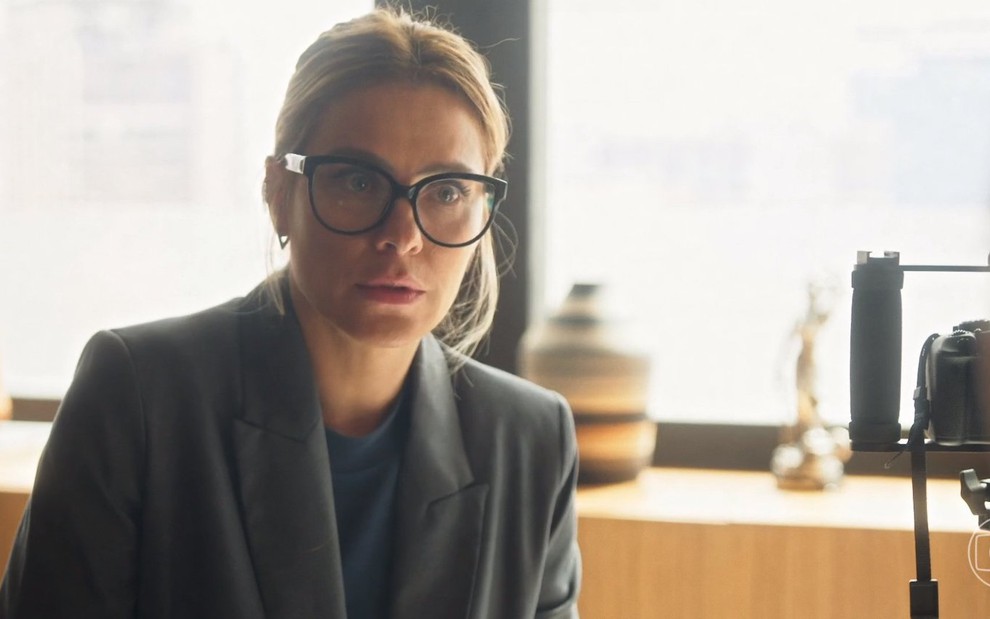 A atriz Carolina Dieckmann está com cabelo preso e usa óculos grandes em cena da novela Vai na Fé como Lumiar