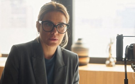 A atriz Carolina Dieckmann está com cabelo preso e usa óculos grandes em cena da novela Vai na Fé como Lumiar