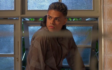 Hugo (MC Cabelinho) sentado em uma janela basculante com expressão séria em cena de Vai na Fé