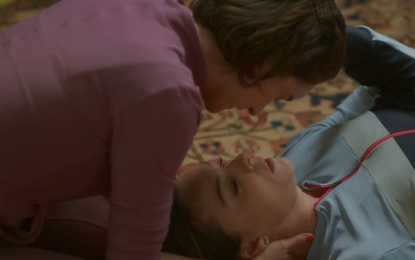 Regiane Alves está deitada no chão, usando um agasalho azul; Priscila Sztejnman se debruça sobre ela, vestindo um casaco rosa, em cena de Vai na Fé