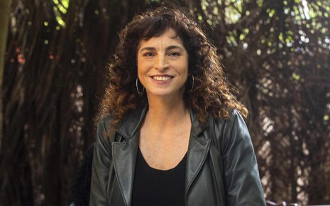 A autora Rosane Svartman usa blusa e jaqueta escuras e está com cabelos cacheados soltos em cena de Cara e Coragem