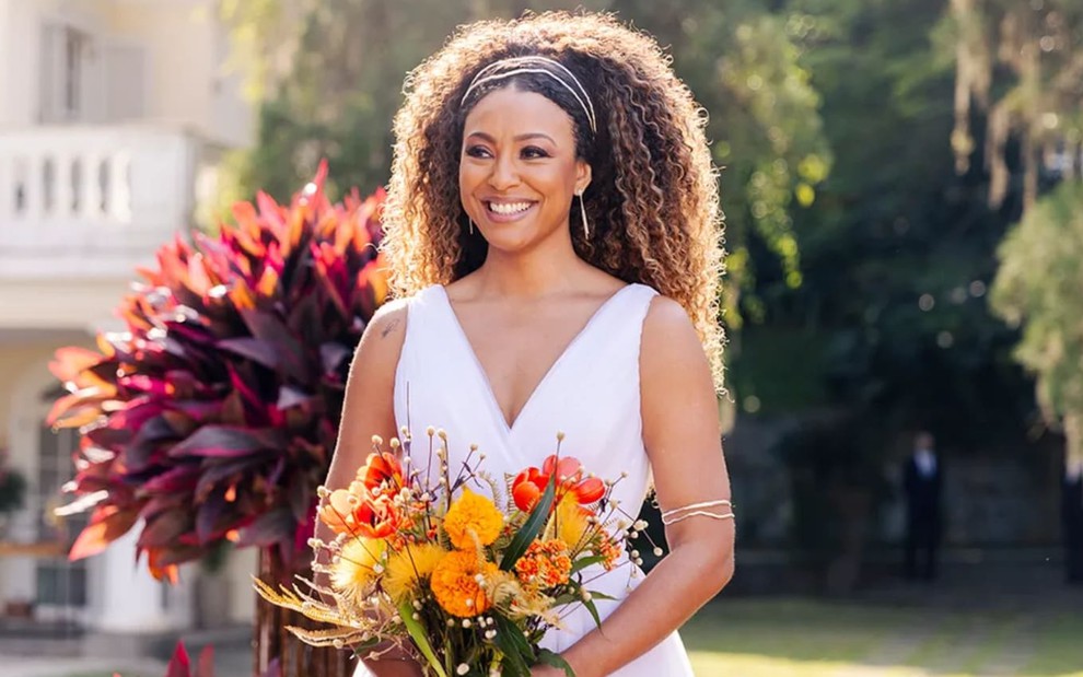 A atriz Sheron Menezzes com vestido de noiva, buqueê de flores, sorrindo em cena de Vai na Fé