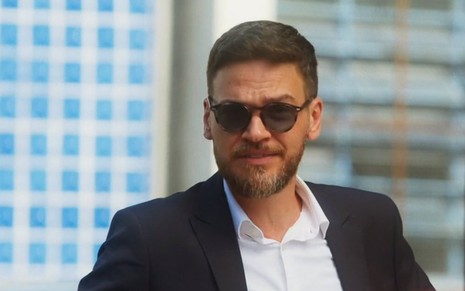 O ator Emilio Dantas usa óculos escuros e camisa branca com terno escuro em cena de Vai na Fé como Theo
