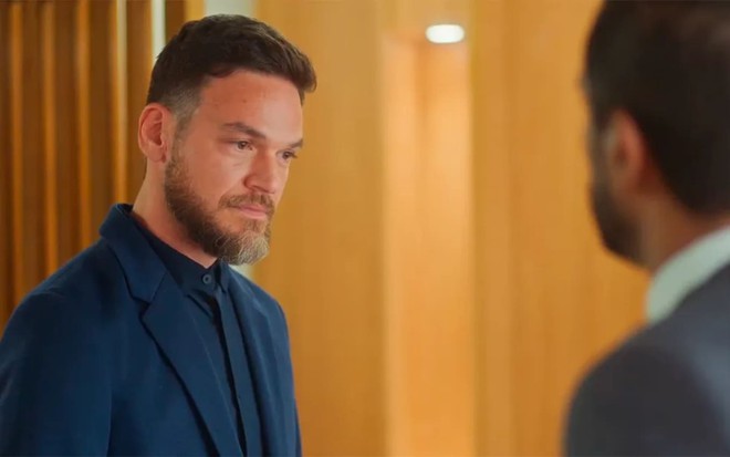 Emilio Dantas caracterizado como Theo em Vai na Fé; ele tem o semblante abalado em cena da novela. Samuel de Assis está de costas para a câmera e encara o colega