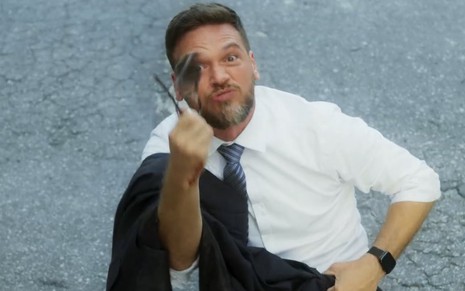 O ator Emilio Dantas está com óculos na mão gesticulando e fazendo careta em cena da novela Vai na Fé