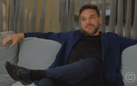 O ator Emilio Dantas está sentado em um sofá em cena da novela Vai na Fé como o vilão Theo