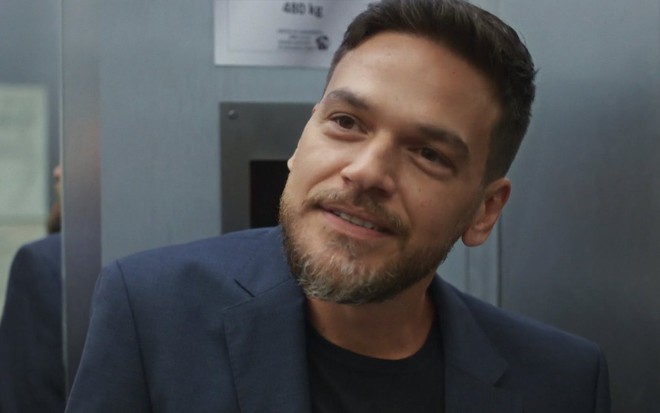 O ator Emilio Dantas está de terno azul e camiseta preta em cena de Vai na Fé caracterizado como Theo
