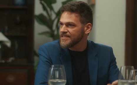 O ator Emilio Dantas usa terno azul, camisa preta e faz expressão de bravo olhando para o lado em cena de Vai na Fé
