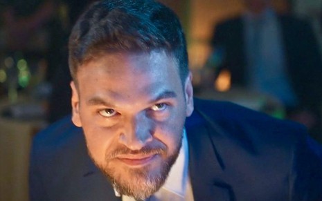 O ator Emilio Dantas está olhando para cima com cara de desgosto em cena de Vai na Fé na qual usa terno e gravata