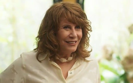 Renata Sorrah caracterizada como Wilma; ele tem os cabelos ruivos na altura dos ombros, usa uma blusa branca e sorri em cena de Vai na Fé