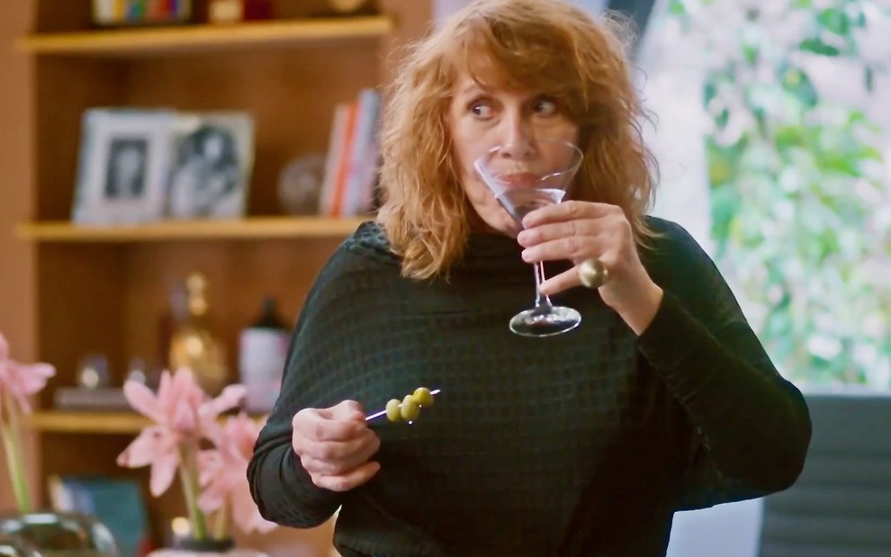 Renata Sorrah leva à boca uma taça com drinque cenográfico em cena de Vai na Fé como a personagem Wilma