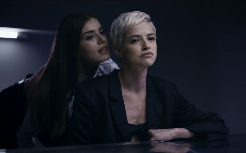 Camila Queiroz grava cena falando no ouvido de Agatha Moreira, que aparece com a expressão séria, como Angel e Giovanna