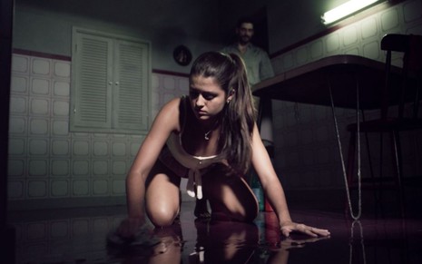 Cena da novela Verdades Secretas 2, do Globoplay, em que Lara (Júlia Byrro) aparece limpando o chão e Nicolau (Julio Machado) surge ao fundo atrás dela sem ela perceber