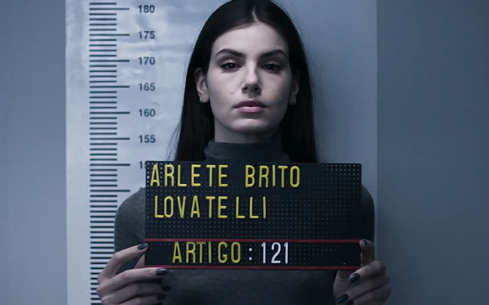 A atriz Camila Queiroz como Angel tira uma foto para registrar a sua ficha criminal segurando uma placa com os dizeres Arlete Brito Lovatelli, artigo: 121 em Verdades Secretas 2