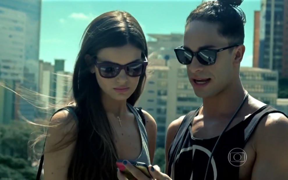 Camila Queiroz grava ao lado de Rainer Cadete, ambos de óculos escuros e olhando o celular, como Angel e Visky de Verdades Secretas