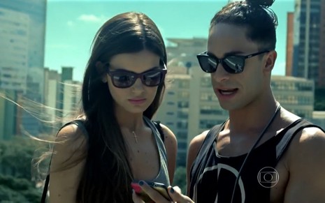 Camila Queiroz grava ao lado de Rainer Cadete, ambos de óculos escuros e olhando o celular, como Angel e Visky de Verdades Secretas