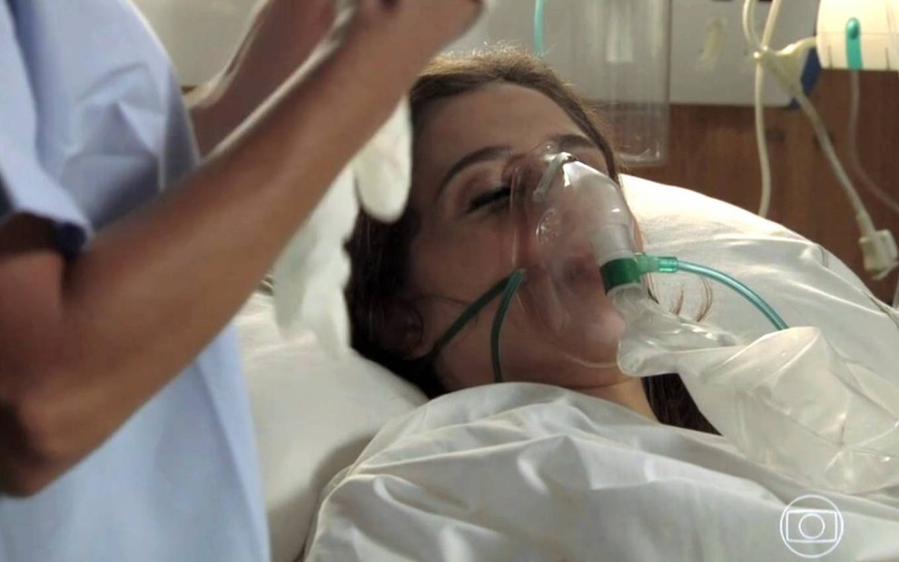 Atriz Marjorie Estiano surge deitada em cama de hospital, com um aparelho para respirara, em cena da novela Império