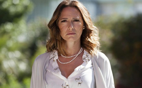 Fernanda Serrano usa camisa social branca e colar de pérolas; ela encara a câmera, série, em cena de A Impostora