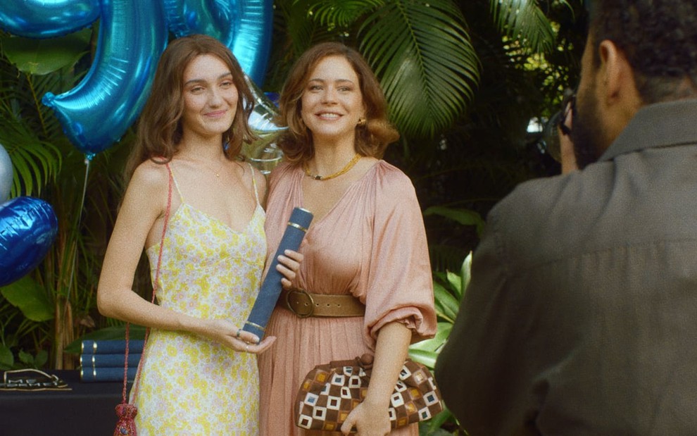 Nina Tomsic, a Liz, usa um vestido verde estampado e segura um canudo de diploma; Leandra Leal, a Tereza, usa um vestido rosa claro. Ambas sorriem para a câmera em cena de A Vida pela Frente