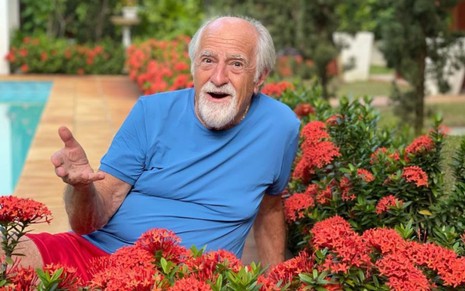 Ary Fontoura veste uma camisa azul e está sorridente; ele está sentado em meio a uma série de flores vermelhas. Ao fundo, é possível ver uma piscina