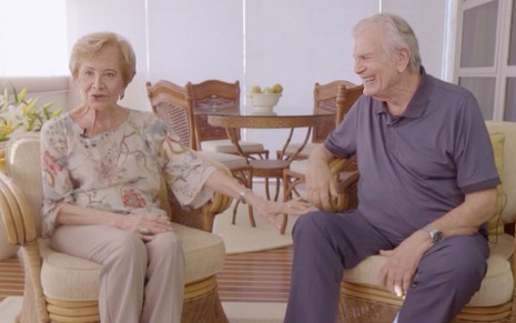 O casal de atores Gloria Menezes e Tarcísio Meira sorri de mãos nadas enquanto eles estão sentados em poltronas na série Os Casais que Amamos, da Globo