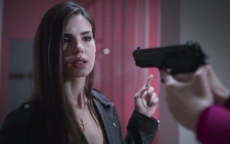 A atriz Camila Queiroz olha para arma apontada para sua cabeça durante gravação de cena de Verdades Secretas 2