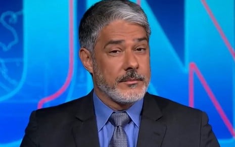 O jornalista William Bonner usa terno e gravata e está no cenário do Jornal Nacional, da Globo