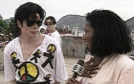 Gloria Maria de frente para Michael Jackson, com microfone na mão, em entrevista de 1996; ele com camiseta do Olodum
