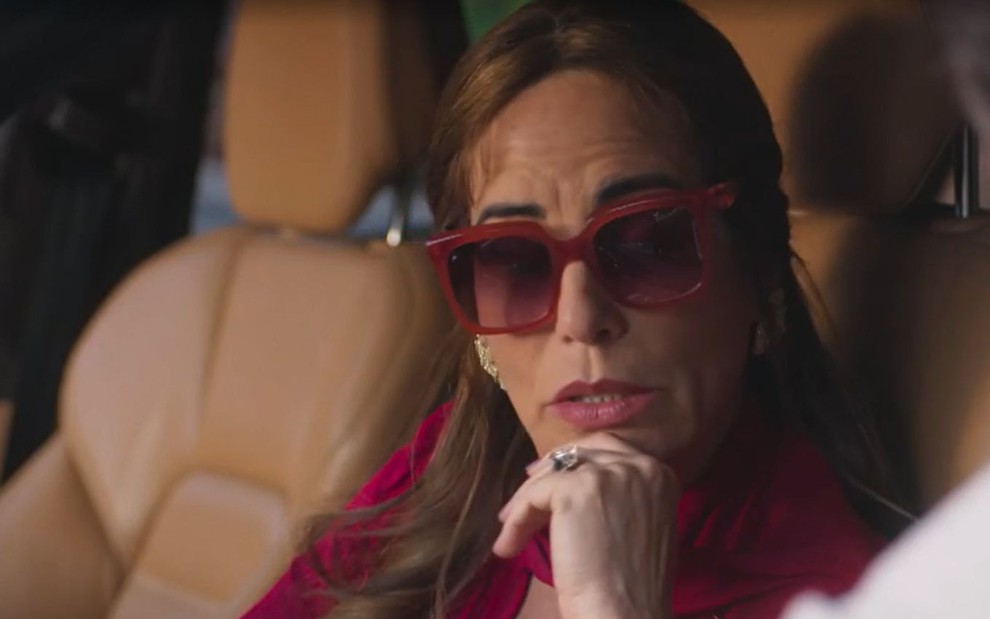 Em cena de Terra e Paixão, Gloria Pires está de óculos escuros dentro de um carro falando com alguém