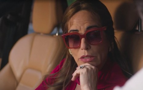 Em cena de Terra e Paixão, Gloria Pires está de óculos escuros dentro de um carro falando com alguém