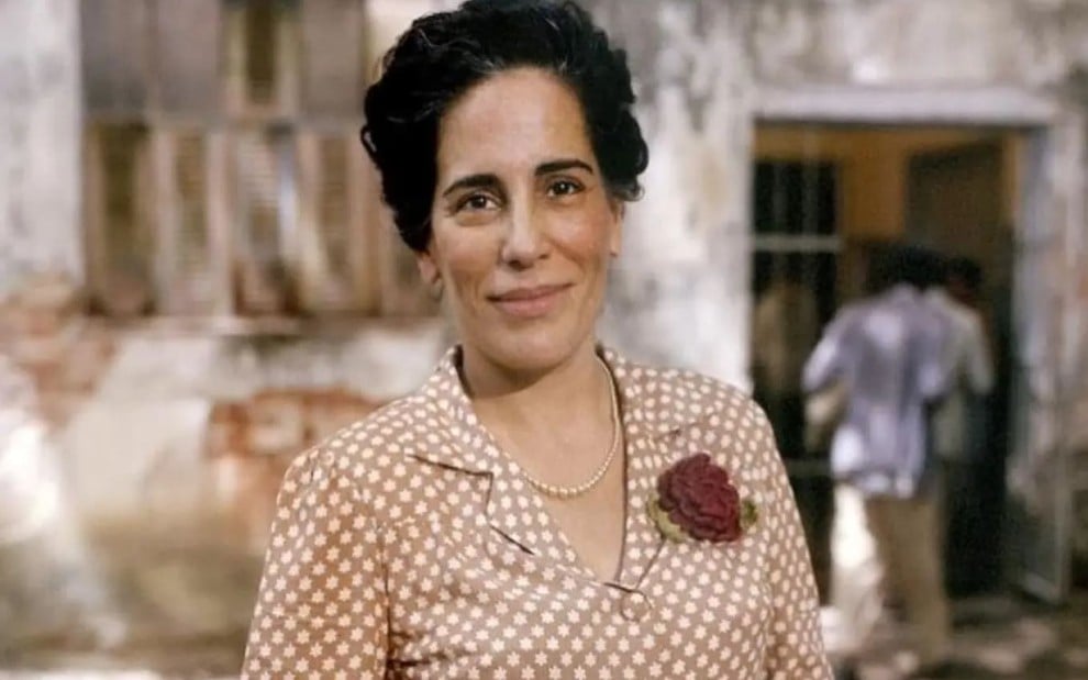 Gloria Pires caracterizada como Nise da Silveira: ela tem os cabelos presos em um coque e veste um terninho bege com estampa poá. Uma rosa está presa na lapela do casaco