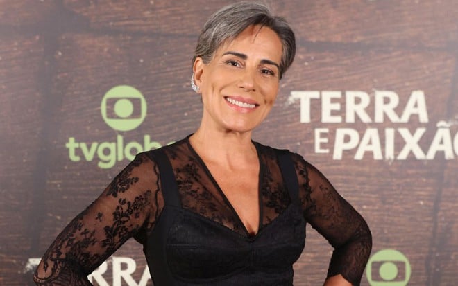 De roupa preta rendada nos braços, Gloria Pires posa com as mãos na cintura no lançamento de Terra e Paixão; atrás há um painel com o nome da novela