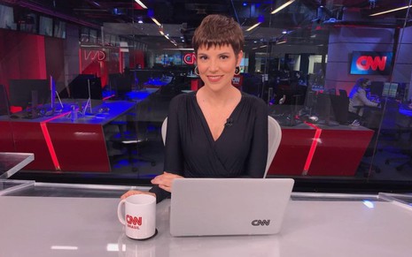 Gloria Vanique com uma blusa preta, sentada na bancada de um telejornal, com uma caneca da CNN Brasil