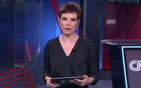 Gloria Vanique com uma blusa escura, segurando um tablet, em um estúdio de telejornal