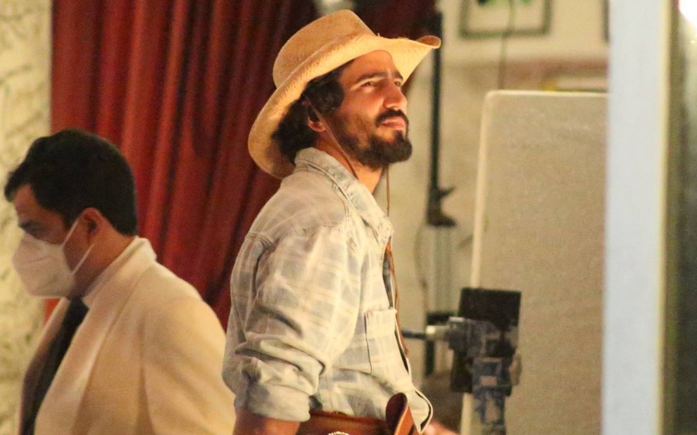 Renato Goés com uma camisa branca quadriculada e um chapéu de sertanejo durante uma gravação de Pantanal, na Globo