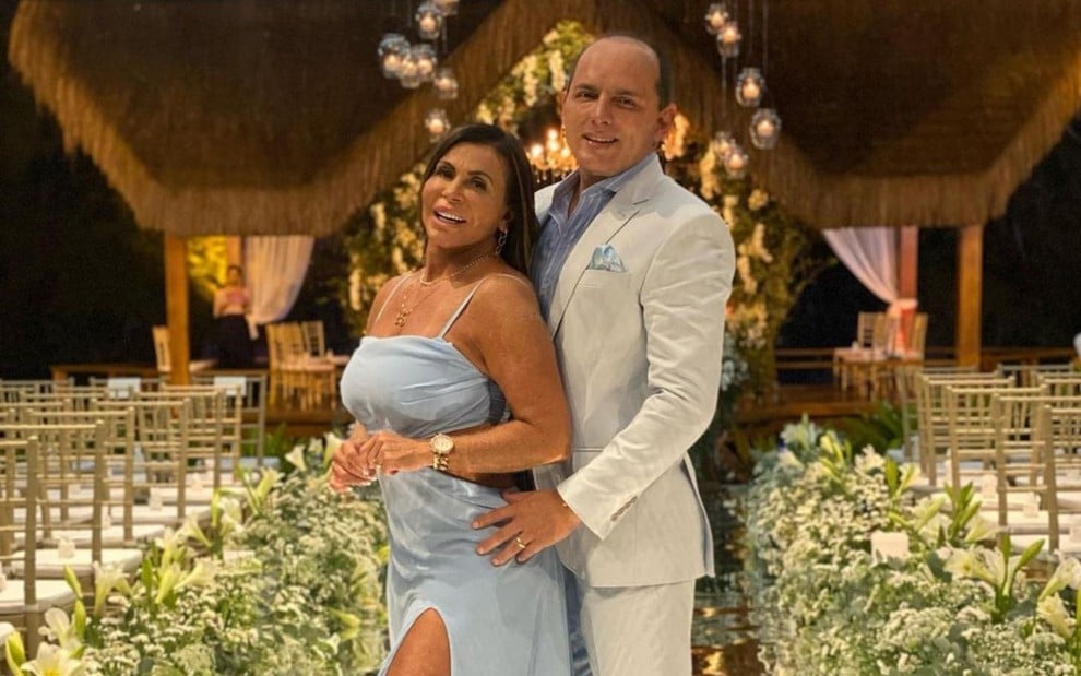 Gretchen veste vestido azul e marido usa terno branco; casal posa abraçado em uma decoração de casamento