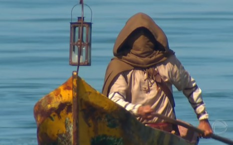 Homem com um capuz marrom tampado o rosto usa um remo em um pequeno barco