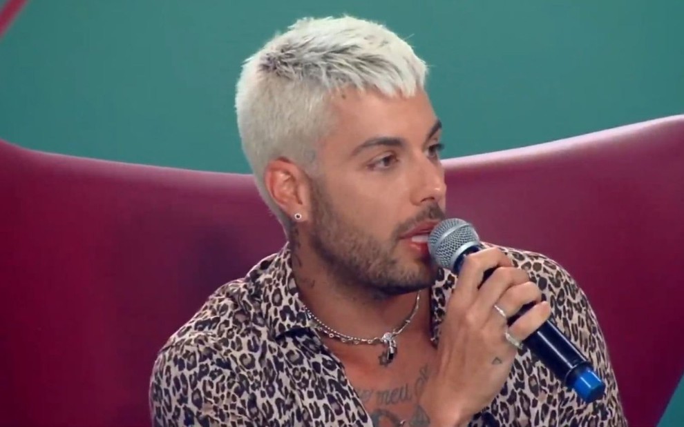 Em cadeira vinho, Gui Araujo usa camisa de botões de oncinha e segura microfone em mãos no programa Hora do Faro, da Record