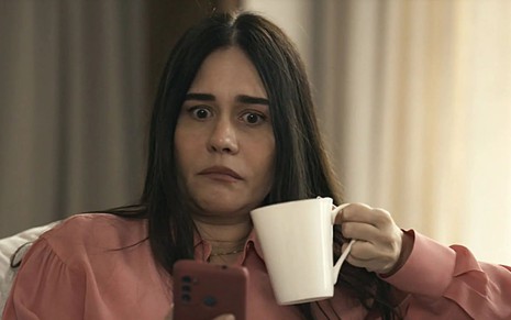 Em cena de Travessia, Alessandra Negrini está olhando o celular, com uma xícara em outra mão; sua expressão é de surpresa