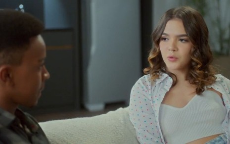 Em cena de Vai na Fé, Mel Maia está conversando com Jean Paulo Campos; ambos estão sentados em um sofá