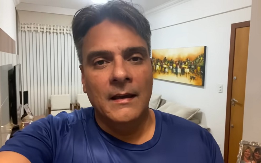 O ex-ator Guilherme de Pádua de camiseta azul, gravando vídeo como selfie, na sala de sua casa