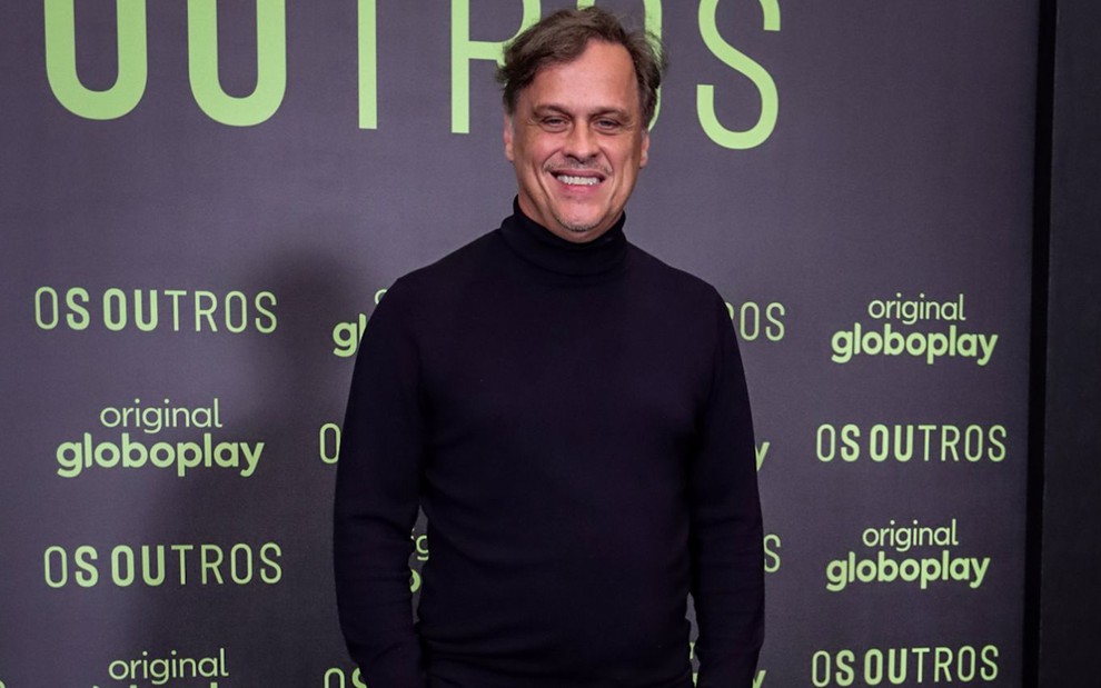Guilherme Fontes posa para foto com uma blusa de gola preta alta; ao fundo está o nome da série Os Outros escrito em verde