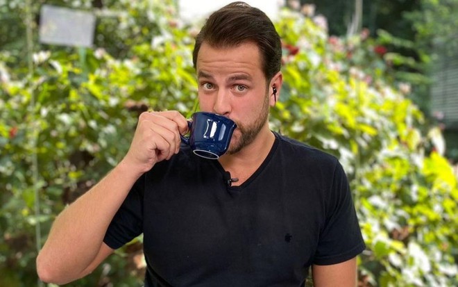 Gustavo Marsengo no Mais Você; ele está sentado, veste uma camiseta preta e bebe de uma xícara de café