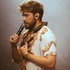 Gustavo Mioto em foto publicada no Instagram; ele está no palco, com um microfone na mão e veste uma camisa branca estampada