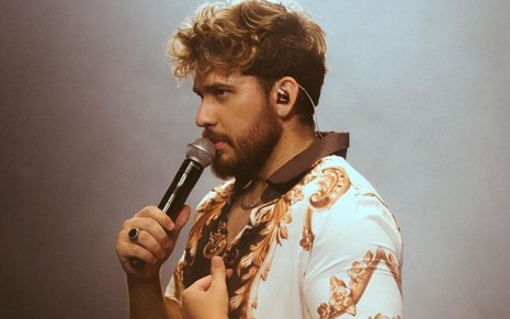Gustavo Mioto em foto publicada no Instagram; ele está no palco, com um microfone na mão e veste uma camisa branca estampada