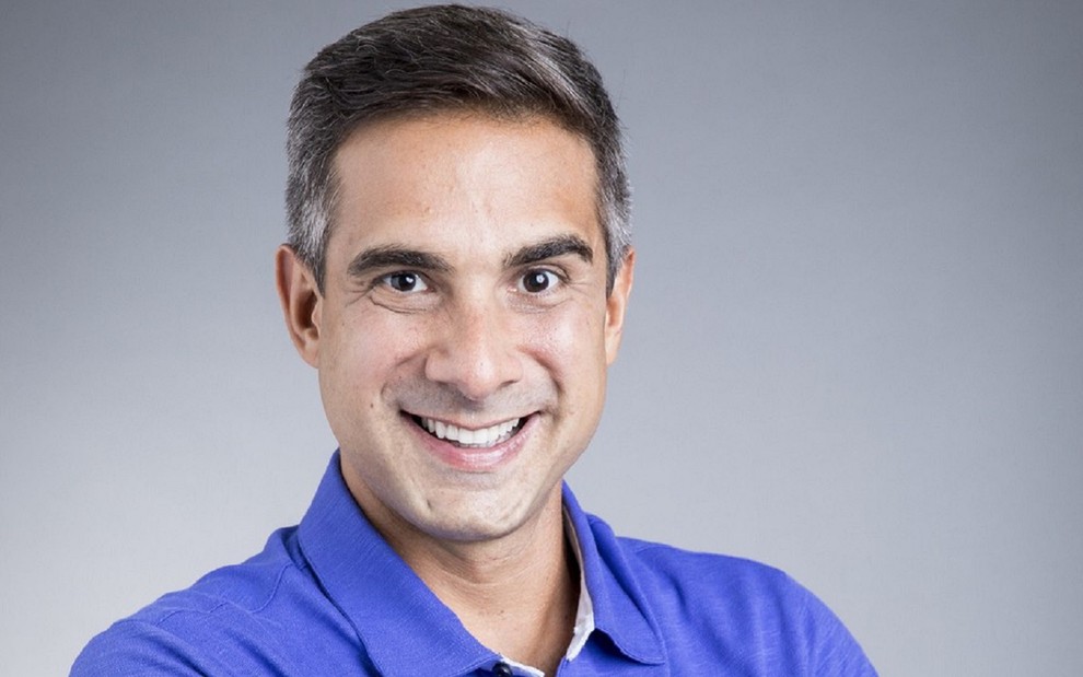 Gustavo Villani sorri e usa uma camisa azul com o logotipo da Globo em um fundo cinza e padrão