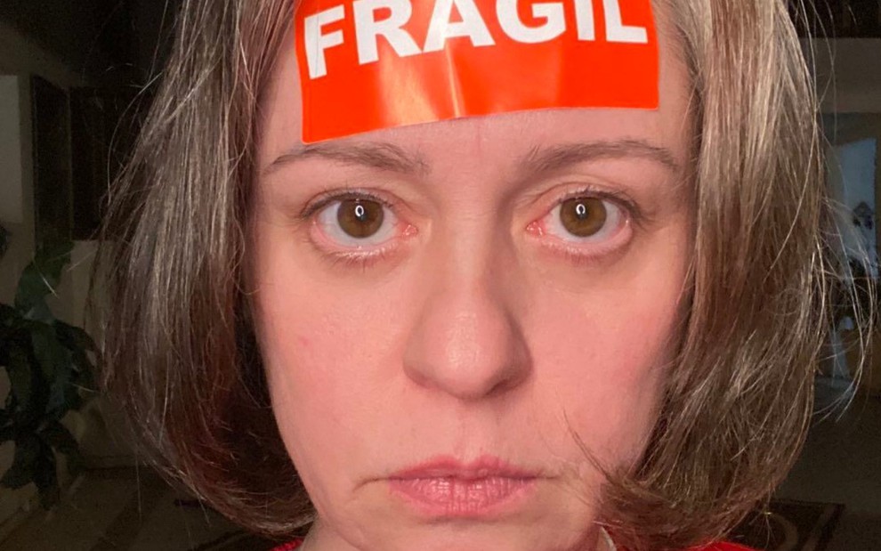 Guta Stresser está com olhar de seriedade e uma fita vermelha com a palavra "frágil" colada em sua testa