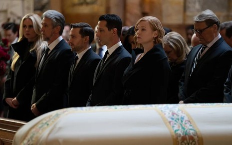 O elenco de Succession está todo de preto em uma igreja, diante de um caixão; os atores têm expressões tristes