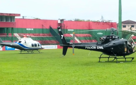 À esquerda, um helicóptero branco está parado em um campo de futebol; à direita, há o helicóptero da Polícia Civil