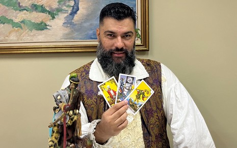 Henrique Zambelli posa segurando cartas ao lado de uma imagem; ele tem barba densa e usa camisa branca com colete